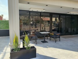 Uma área externa com mesas, cadeiras, janelas amplas e plantas em vasos.