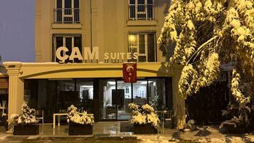 Un hotel con un letrero que dice "CAM SUITES" y una bandera con una estrella. Hay plantas en macetas frente a la entrada.