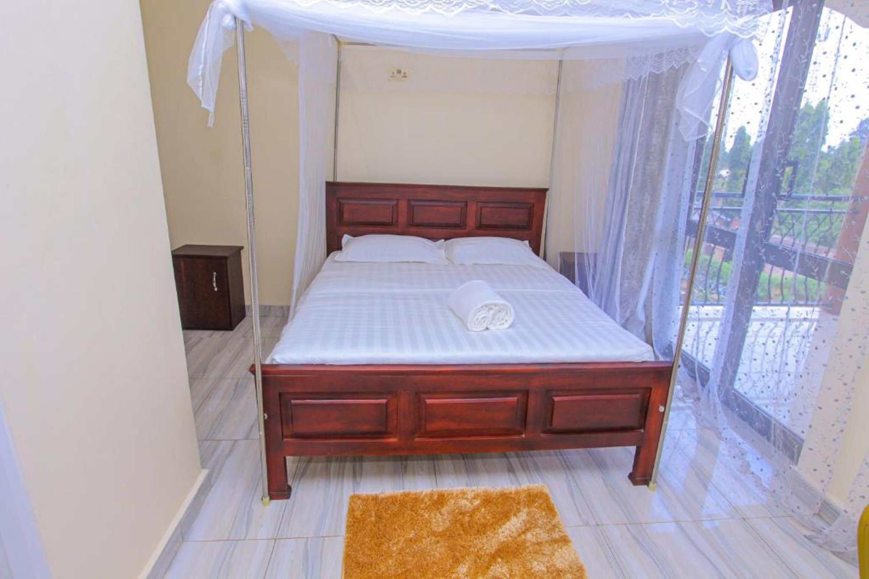A bedroom with a wooden bed, a white canopy, a small wooden cabinet, and a view of greenery outside.