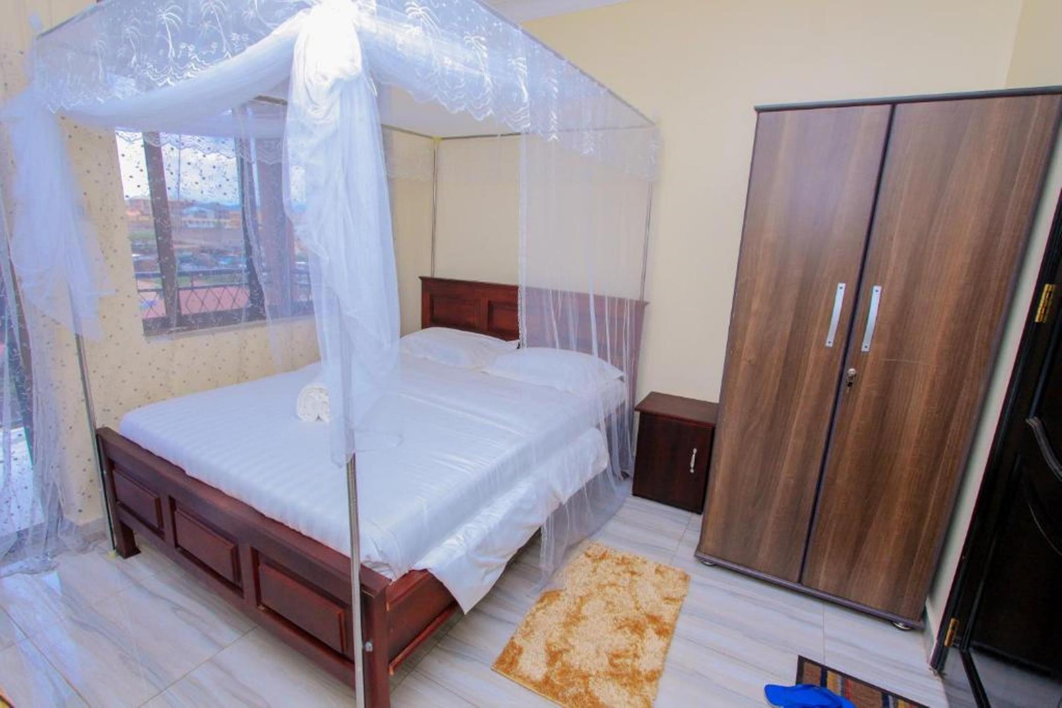 A bedroom with a bed under a mosquito net, a wooden wardrobe, and a small bedside table.