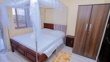 A bedroom with a bed under a mosquito net, a wooden wardrobe, and a small bedside table.