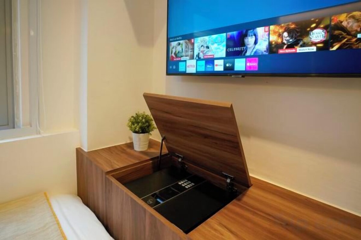 A room with a bed, a wooden bedside table with a plant, and a mounted television displaying various channels.