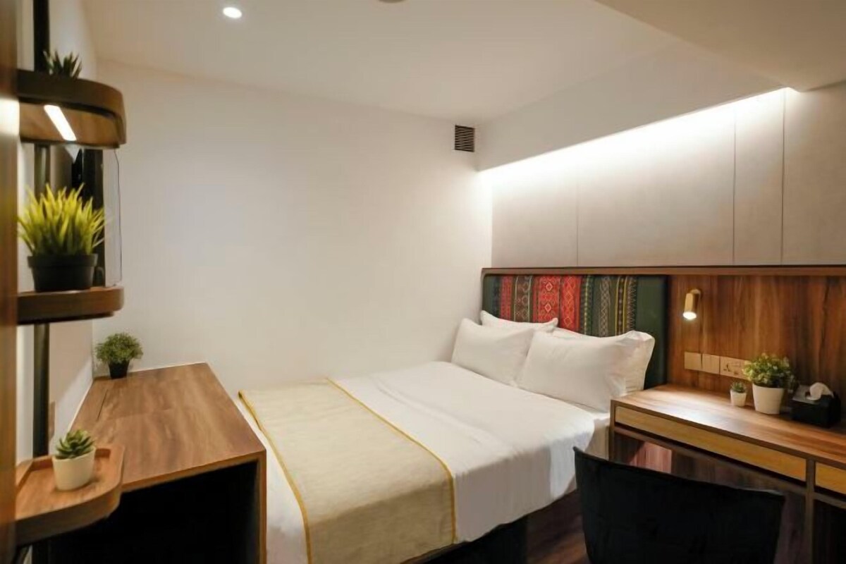 A modern hotel room with a bed, bedside table, and a desk with a chair. There are plants on the bedside table and a shelf with plants on the wall.