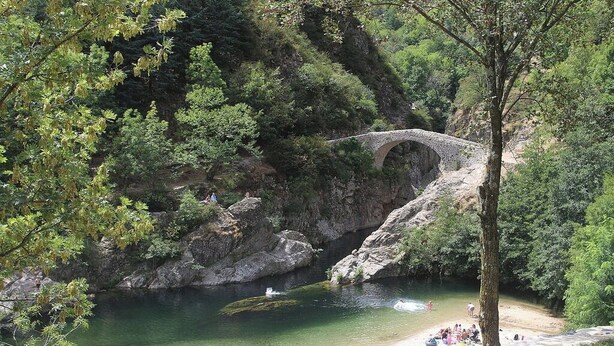 Un pont de pedra sobre un riu, envoltat d'arbres i una platja sorrenca amb gent.