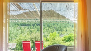 Habitació moderna amb taula de vidre, una butaca gris i vista a un paisatge verd i abundant a través de grans finestres.