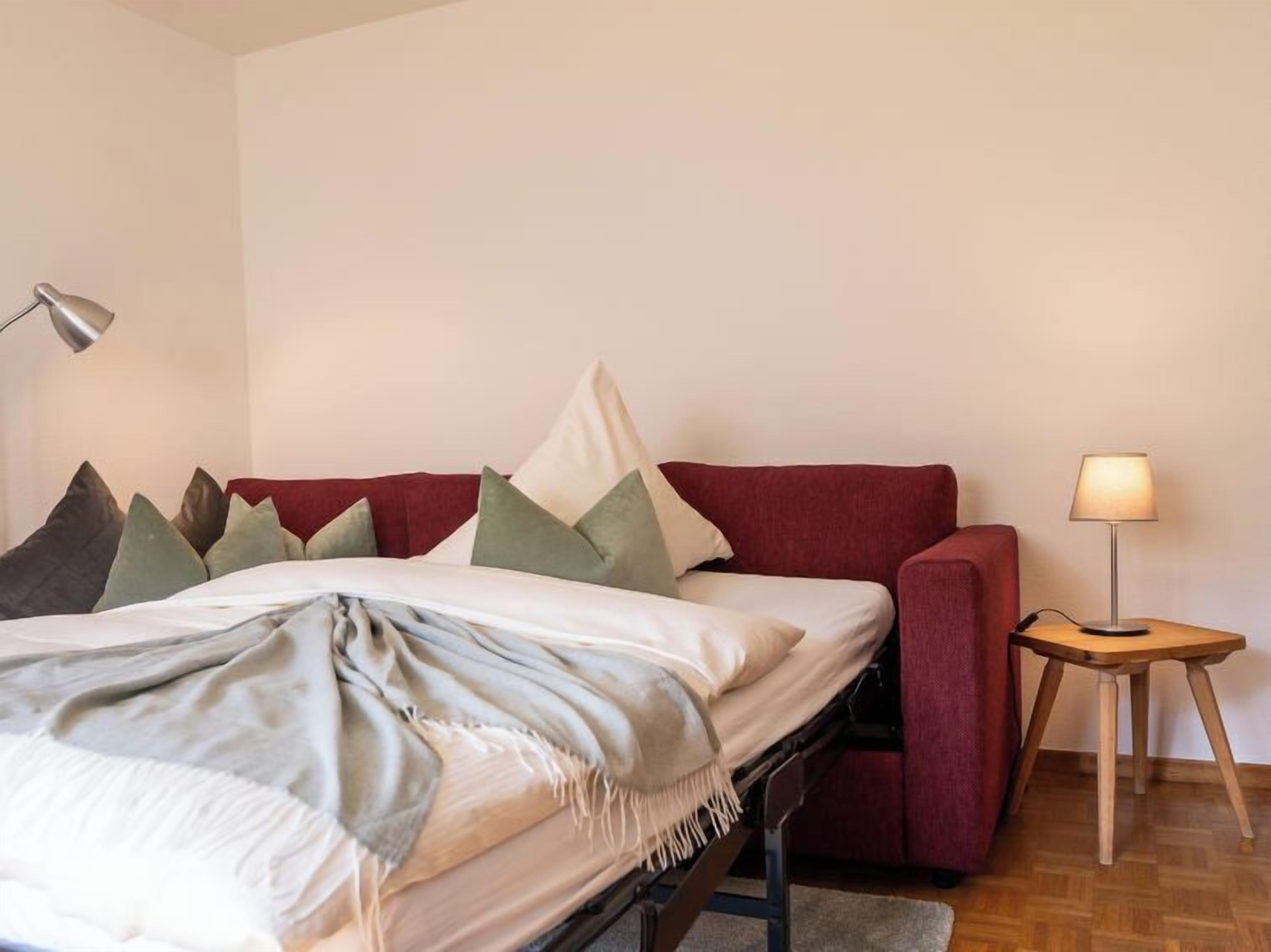 A bedroom with a bed, a red sofa, a small table with a lamp, and a floor lamp.