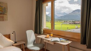 A hotel room with a bed, a desk with a chair, a window with a view of mountains and a green landscape, and a lamp on a bedside table. - Chesa Muntarütsch by Interhome (Samedan)