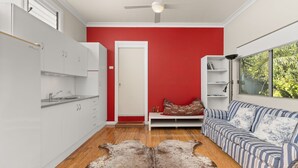 A compact living space with a kitchenette, a red accent wall, a sofa with striped cushions, and a view of greenery through a window. - Kingston Gardens by Experience Jervis Bay (Huskisson)