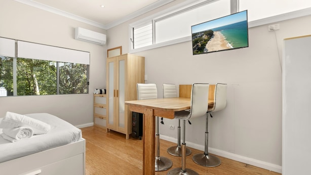 A modern room with a wooden dining table, white chairs, a sofa, and a TV displaying a beach scene. - Kingston Gardens by Experience Jervis Bay (Huskisson)