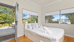 A modern bedroom with a large bed, two windows, and a sliding glass door leading to an outdoor area. - Kingston Gardens by Experience Jervis Bay (Huskisson)