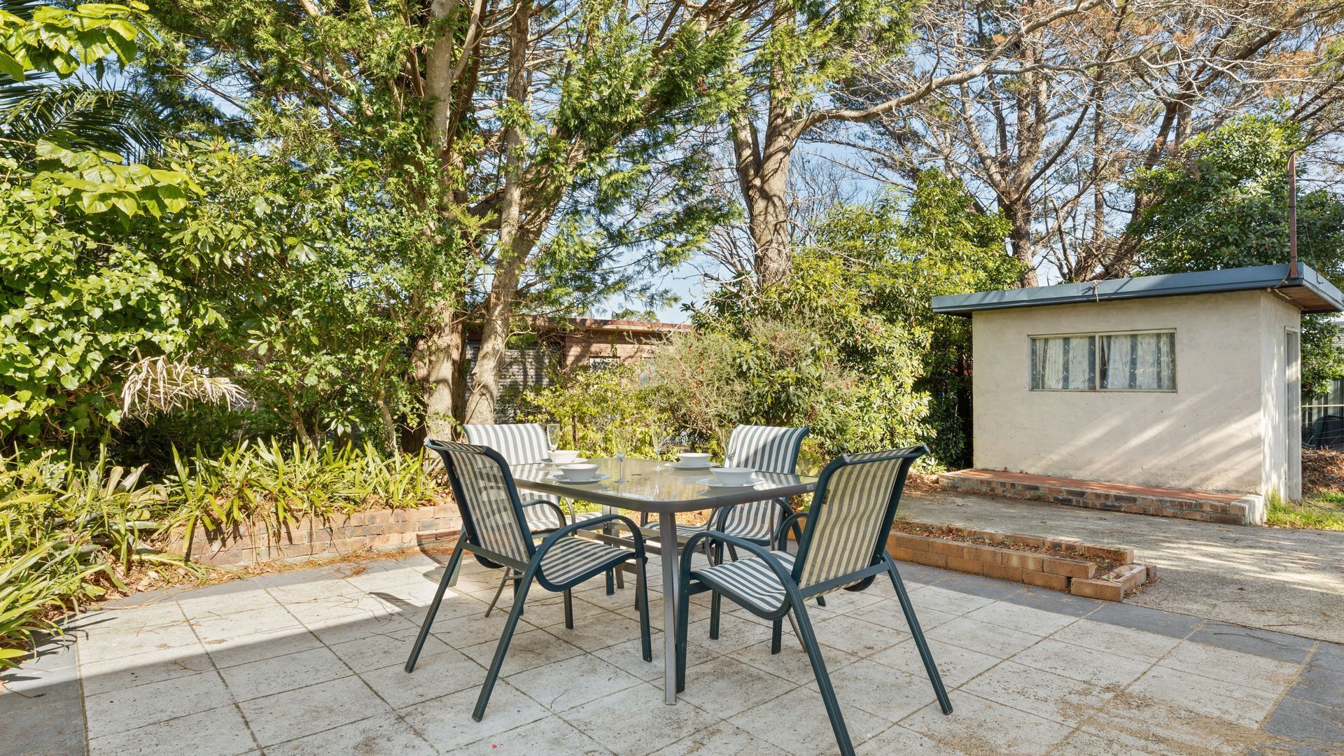 A patio with a round table and chairs, surrounded by lush greenery and trees.