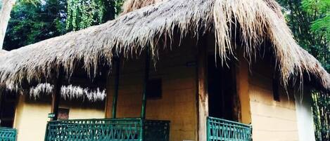 A thatched-roof hut with a wooden door and a small porch with a green railing.