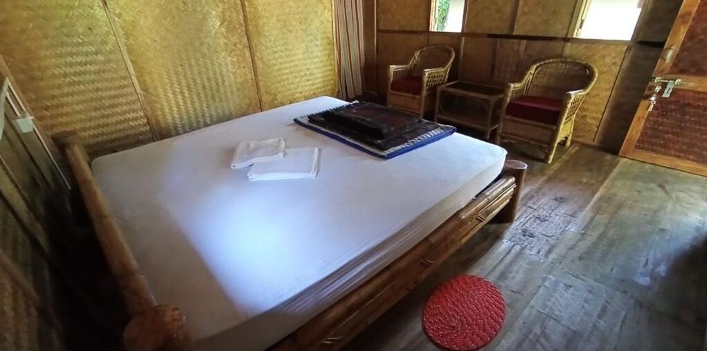 A room with a bed, a red woven stool, and two wicker chairs.