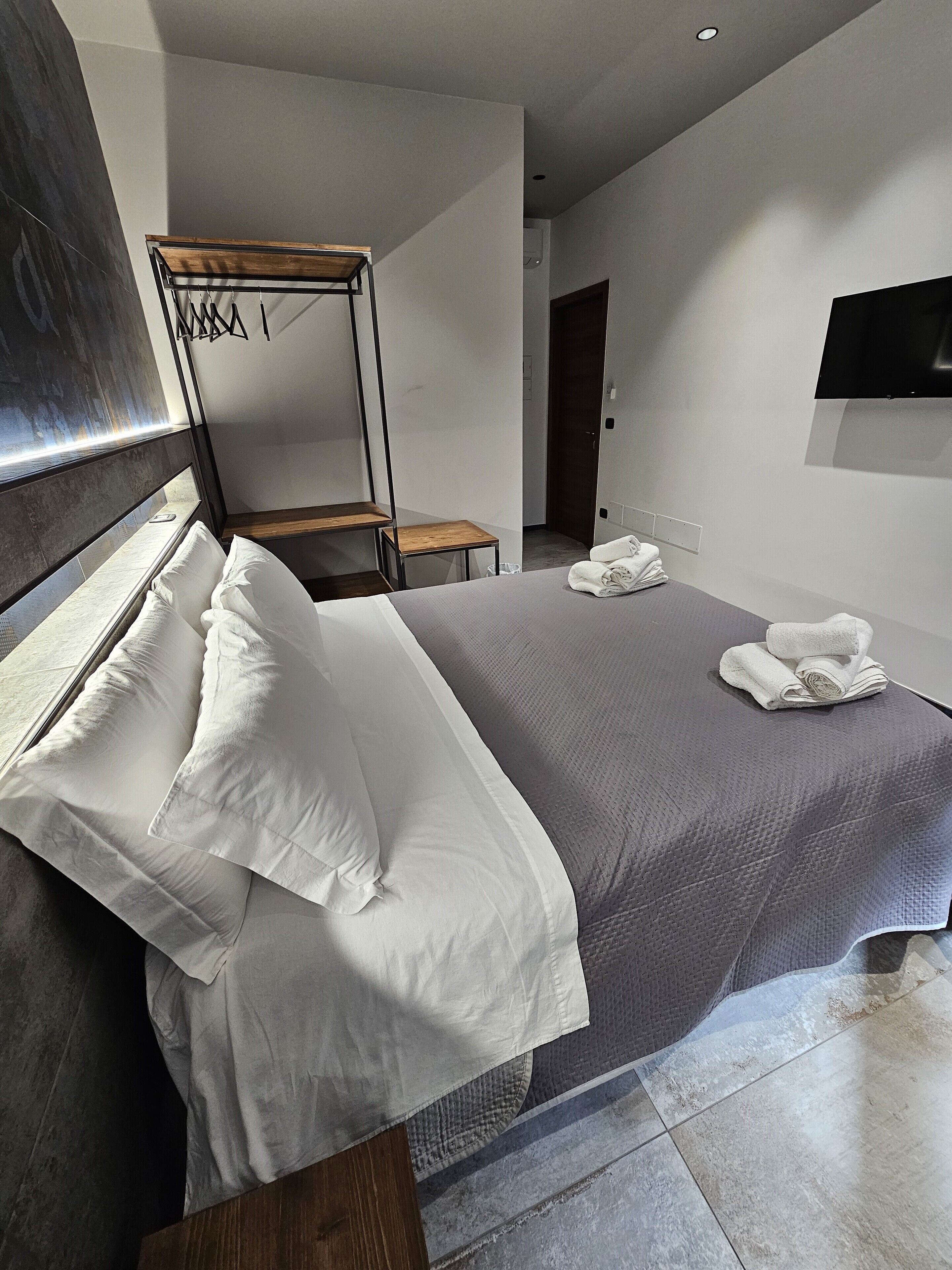 A modern hotel room with a large bed, a wooden bedside table, a small wooden chair, and a flat-screen TV mounted on the wall.