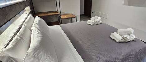 A modern hotel room with a large bed, a wooden bedside table, a small wooden chair, and a flat-screen TV mounted on the wall.