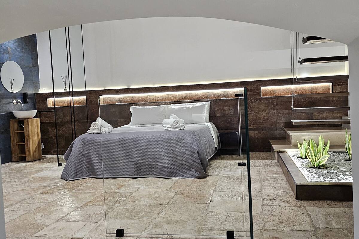 A modern bedroom with a large bed, a built-in bench, a small plant area, and a stone-tiled floor.