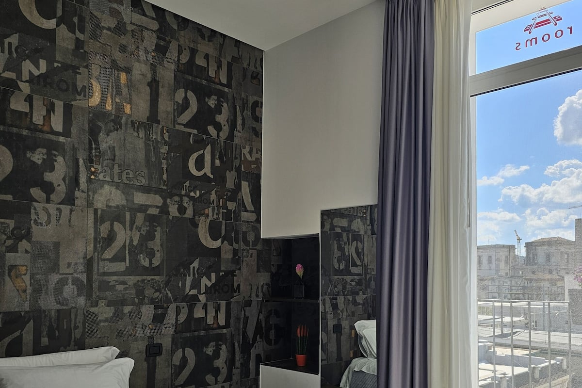 A hotel room with a bed, a desk, and a view of the city through large windows.