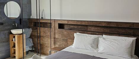 A modern bedroom with a large bed, a wooden headboard, a round mirror, a wooden side table, and a stone wall.