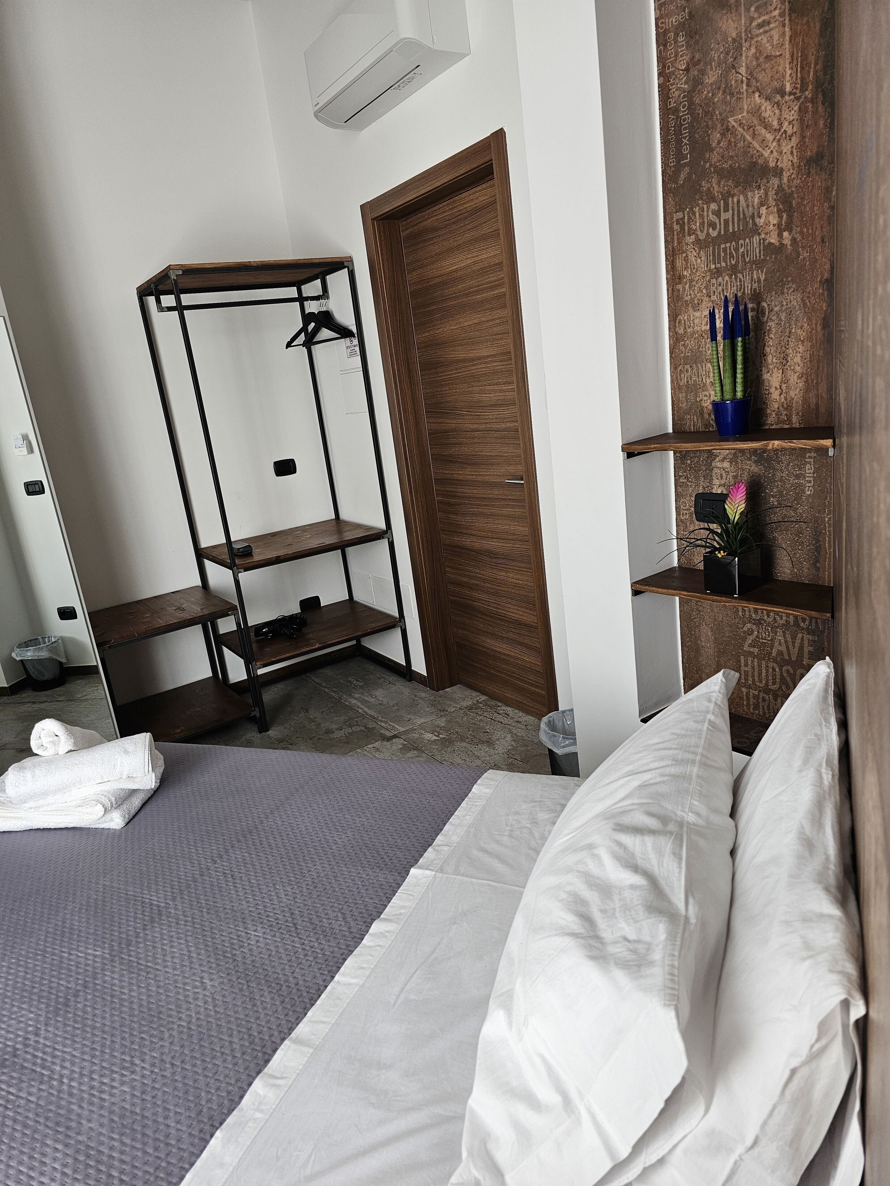 Een netjes ingerichte hotelkamer met een bed, een houten garderobe, een metalen schapkas en een klein plankje met decoratieve objecten.