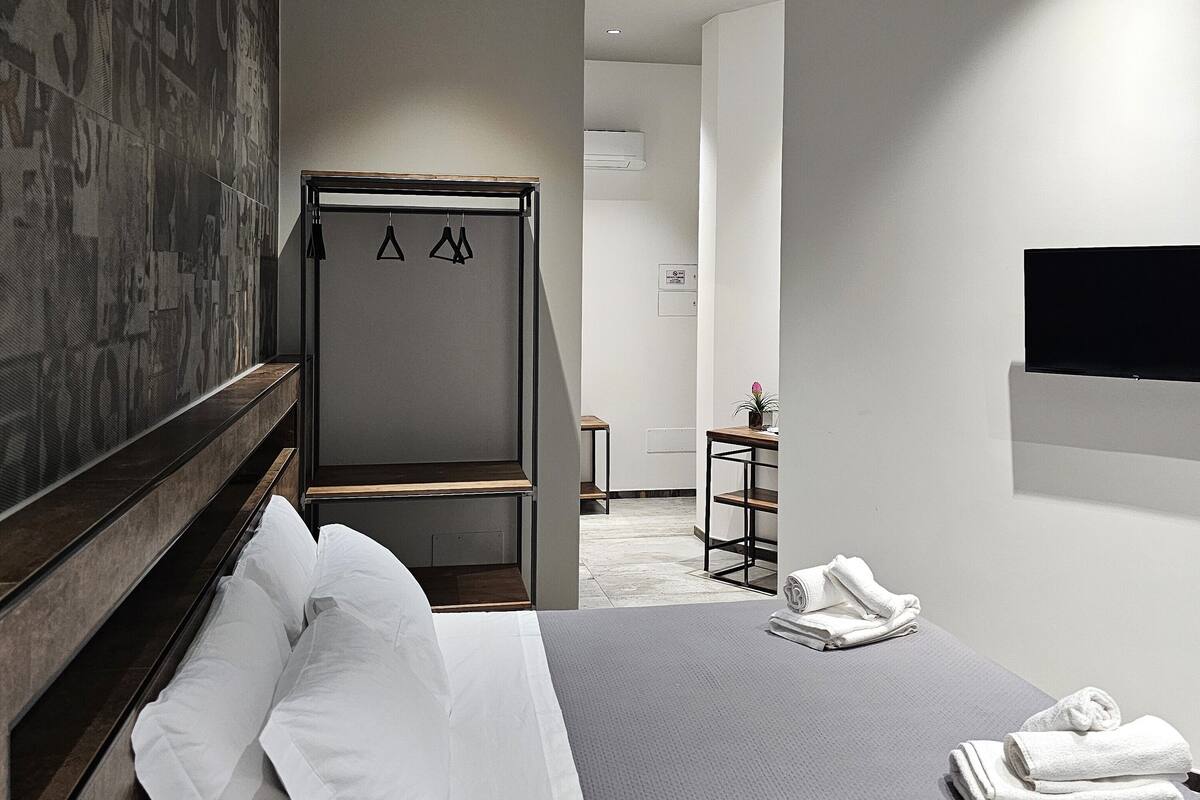 A modern hotel room with a neatly made bed, a small table with a plant, and a flat-screen TV mounted on the wall.