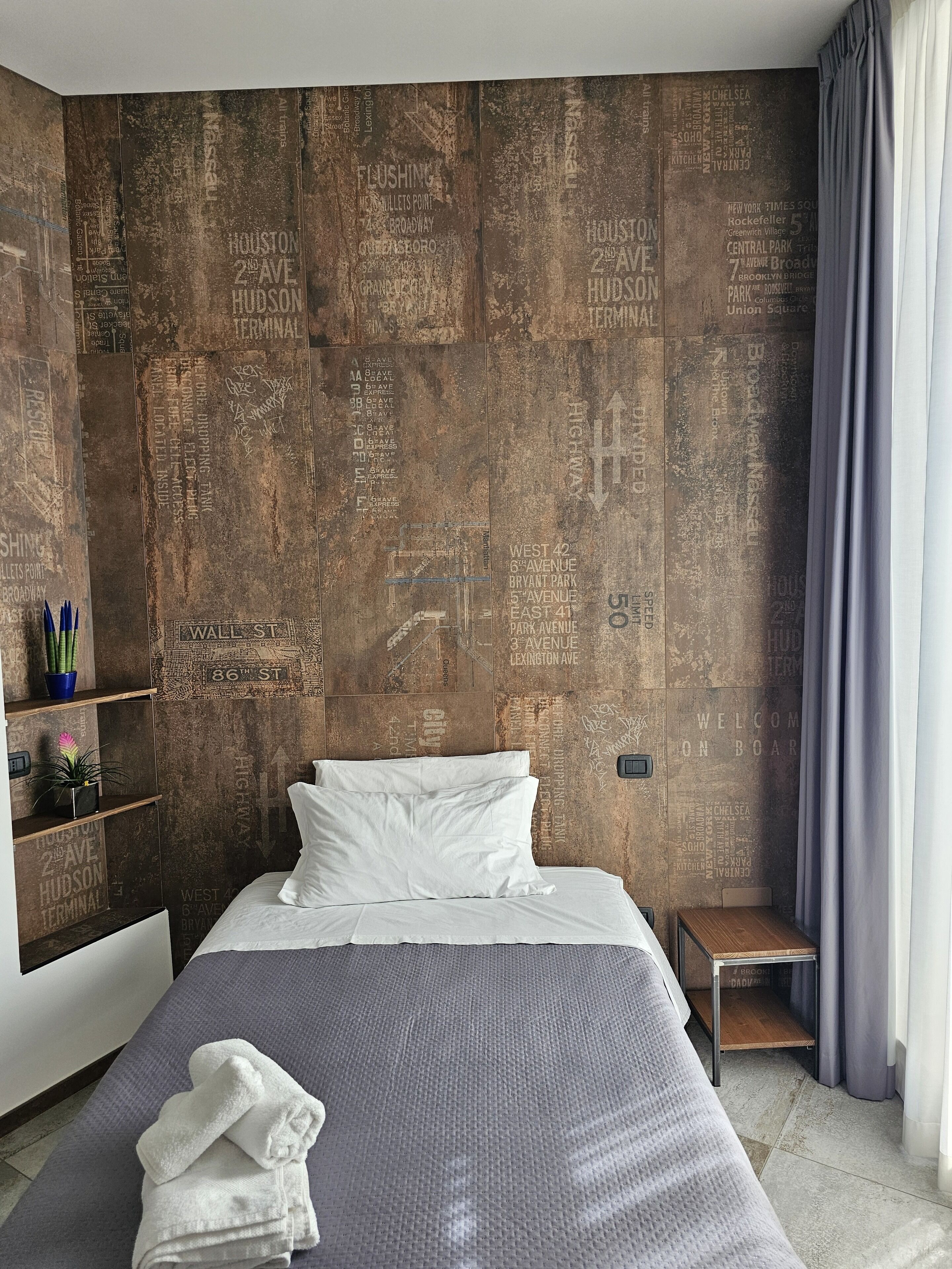 Un dormitorio moderno con pared texturizada, una cama con ropa blanca, una mesita de noche pequeña y una ventana con cortinas.
