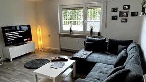 Una sala de estar moderna con un sofá gris, una mesita de centro blanca y un televisor de pantalla plana sobre un mueble blanco. Se ve una ventana con persianas, un radiador y algunas fotografías enmarcadas en la pared.