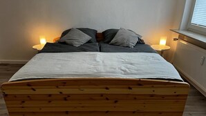 Una cama de madera con colchón blanco y dos almohadas, flanqueada por lámparas de noche, en una habitación con radiador y ventana.