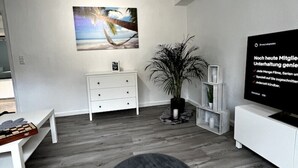 Una sala de estar moderna con un sofá blanco, un reposapiés negro, un televisor de pantalla plana, un cuadro grande y una planta en maceta.
