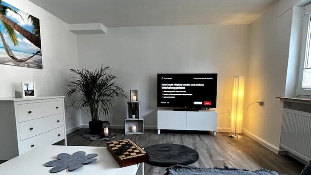 Una sala de estar moderna con un televisor de pantalla plana, un mueble blanco, una planta, un tablero de ajedrez y un rincón de descanso acogedor.