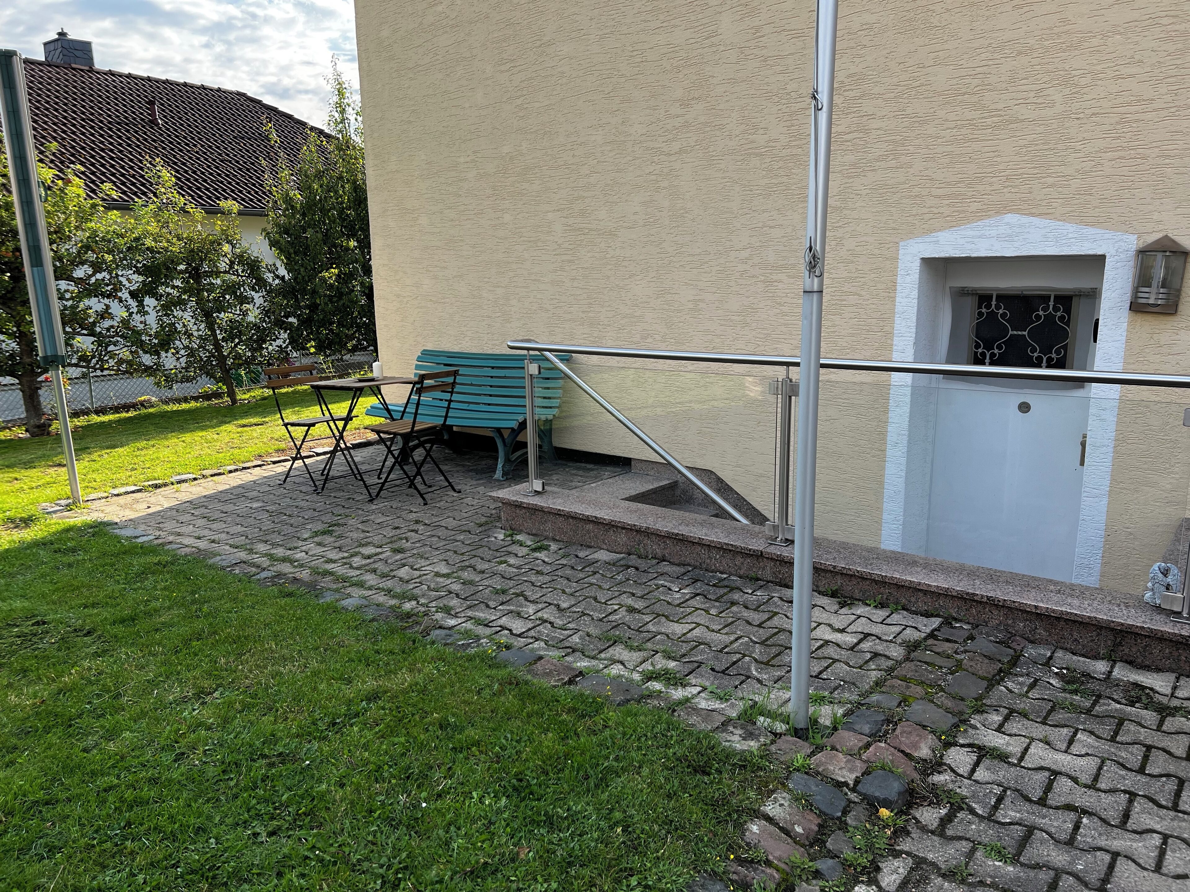 A paved outdoor area with a bench and a table, a metal railing, and a building entrance with a door and window.