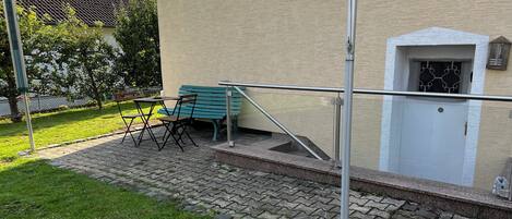 A paved outdoor area with a bench and a table, a metal railing, and a building entrance with a door and window.