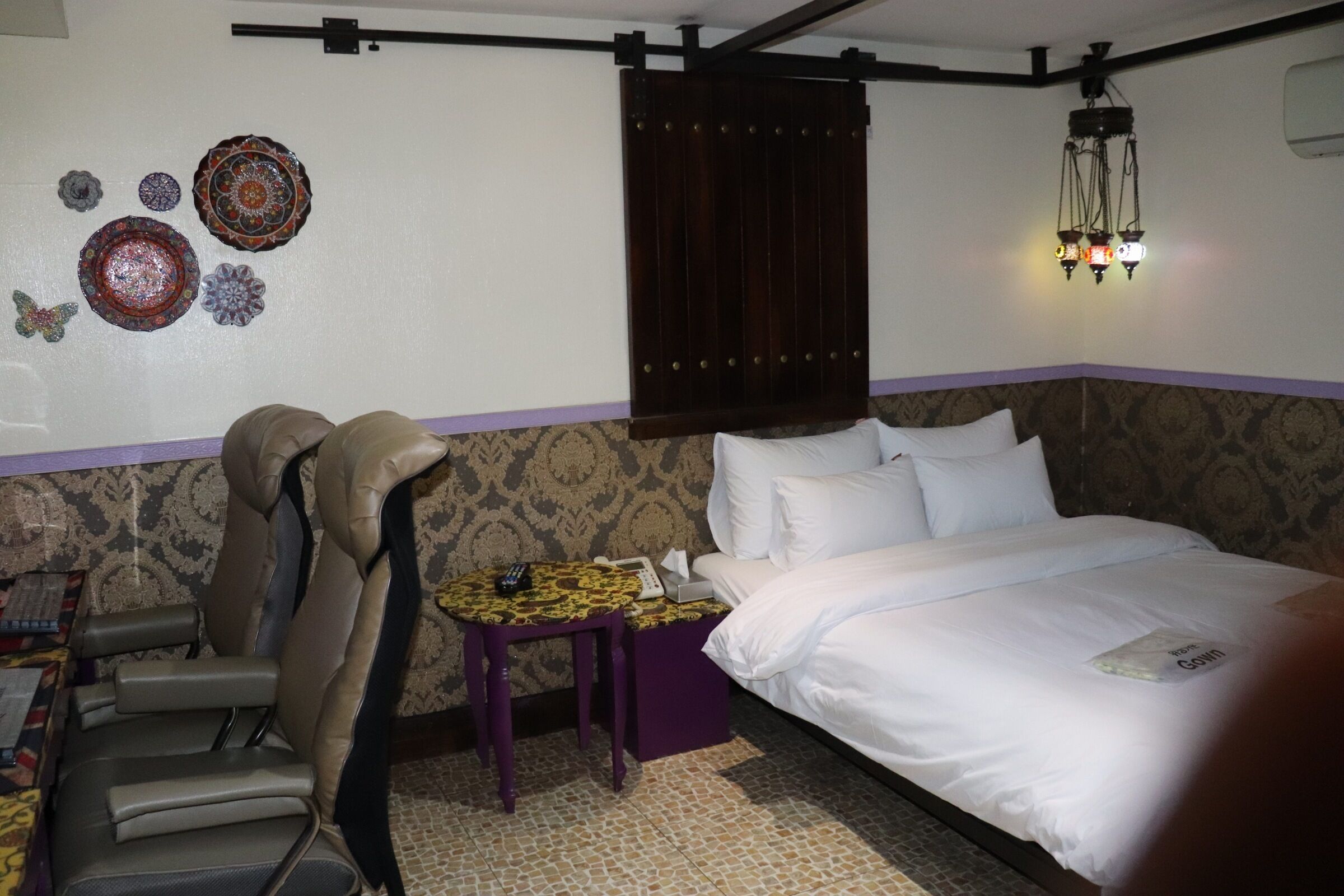A hotel room with a bed, a chair, a small table with a phone and remote, decorative plates on the wall, and a hanging light fixture.
