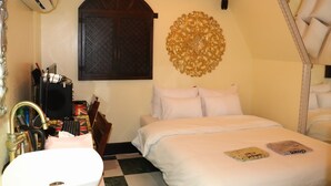 Ein Hotelzimmer mit Bett, Wanddeko, einem Fenster mit dunklem Holztür und einem Schreibtisch mit Computer und diversen Gegenständen.