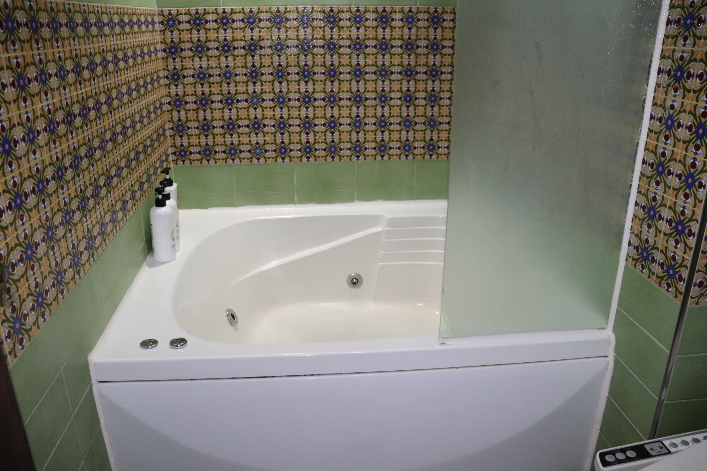 Casa de banho com banheira branca, paredes com azulejos verdes e painel de azulejos estampados.
