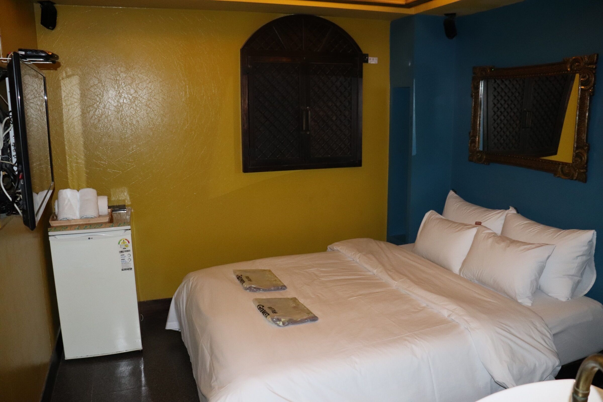 A hotel room with a bed, a small refrigerator, a window with a decorative grille, and a mirror with an ornate frame.