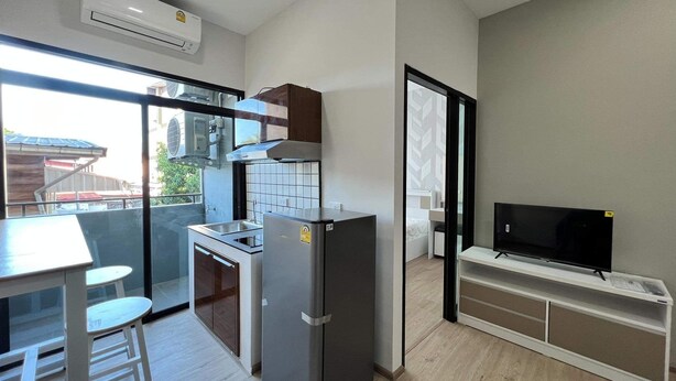 A compact kitchen with a refrigerator, stove, and sink. There's a dining area with a table and chairs, and a TV on a stand. - Phattharasaya Home (Udon Thani)