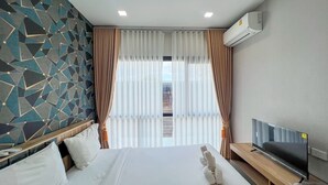 A hotel room with a geometric-patterned wall, a bed with white linens, a TV on a wooden stand, and an air conditioning unit on the wall. - Phattharasaya Home (Udon Thani)