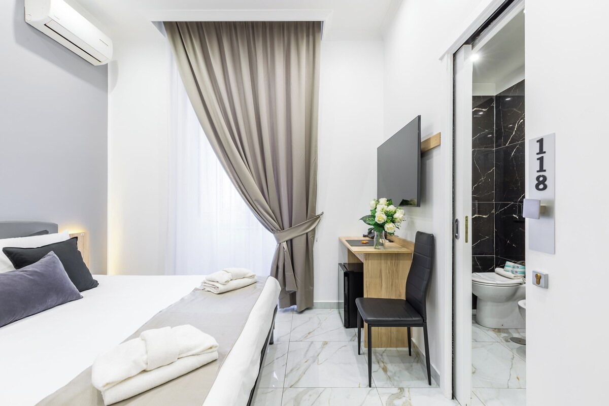 A modern hotel room with a bed, desk, chair, and a bathroom with a toilet and sink.