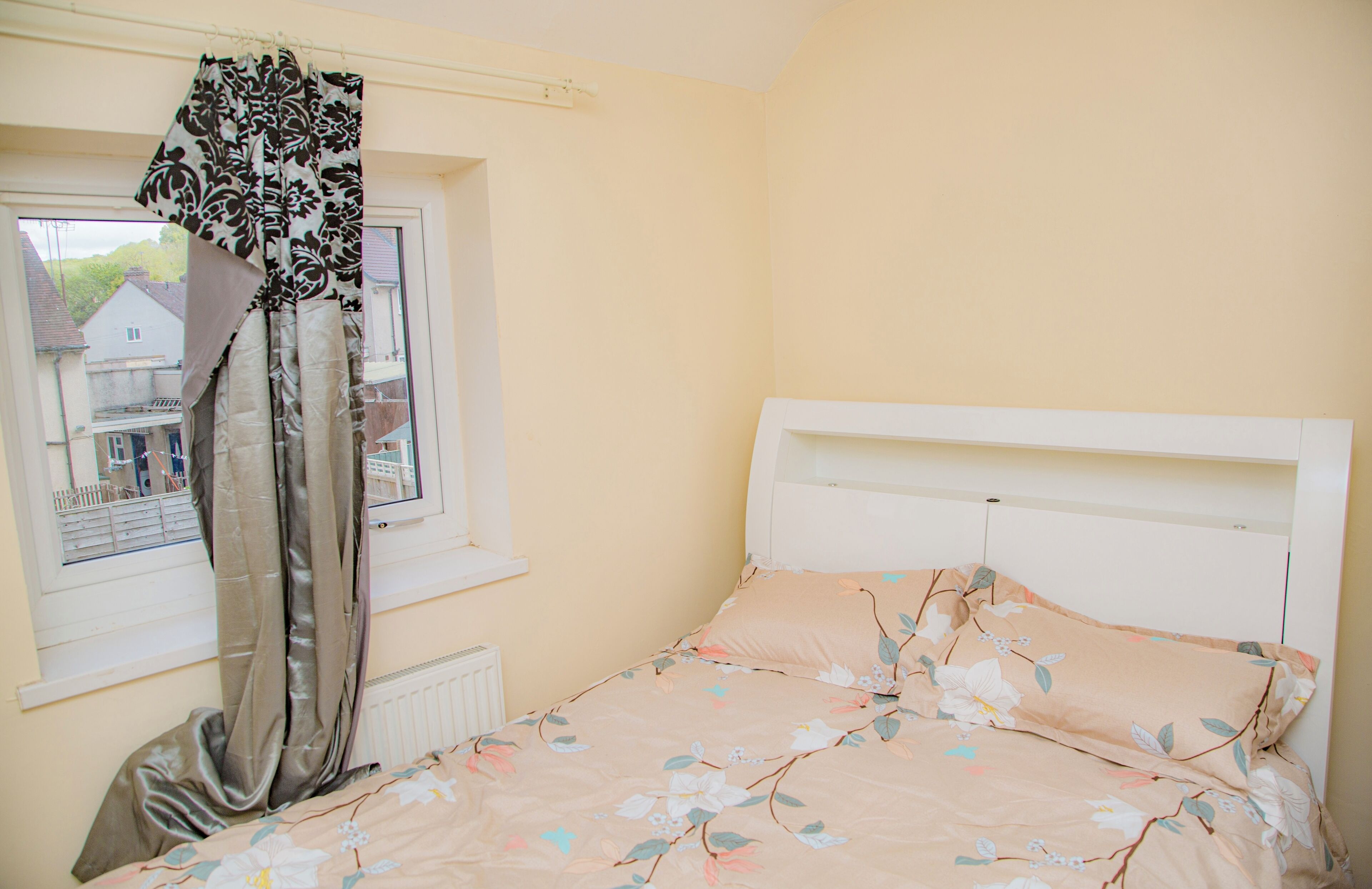 A bedroom with a bed, a window with curtains, and a view of houses outside.