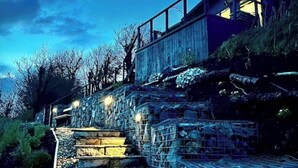 Eine Treppe führt zu einer Holzhütte mit einem Glasbalkon, umgeben von Steinmauern und Vegetation.