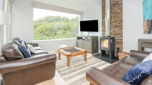 A modern living room with a fireplace, leather sofas, a flat-screen TV, and a large window overlooking a forested area. - 9 Cedars (St. Agnes)