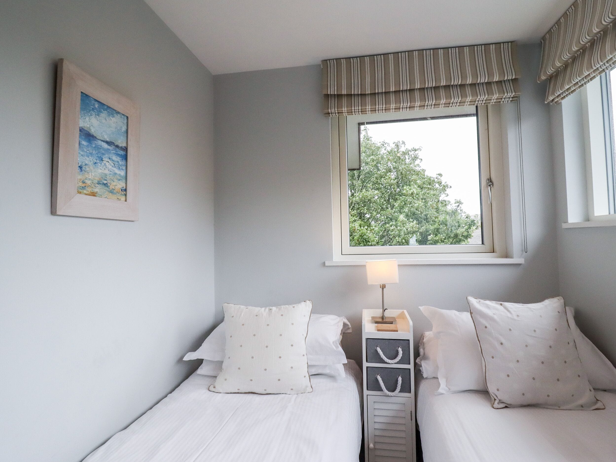 A bedroom with two beds, a window with curtains, a bedside lamp, and a painting on the wall.