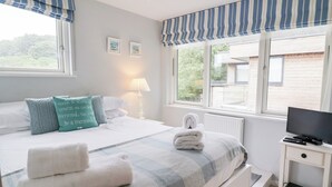 A neatly made bed with pillows, a bedside lamp, a TV on a small table, and a window with curtains. - 9 Cedars (St. Agnes)