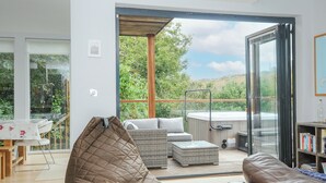 A modern living room with a large sliding glass door leading to a balcony, wicker furniture, and a hot tub. - 9 Cedars (St. Agnes)