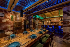 Un restaurante con una larga mesa de madera lista para comer, paredes de ladrillo y una zona de bar con iluminación azul.