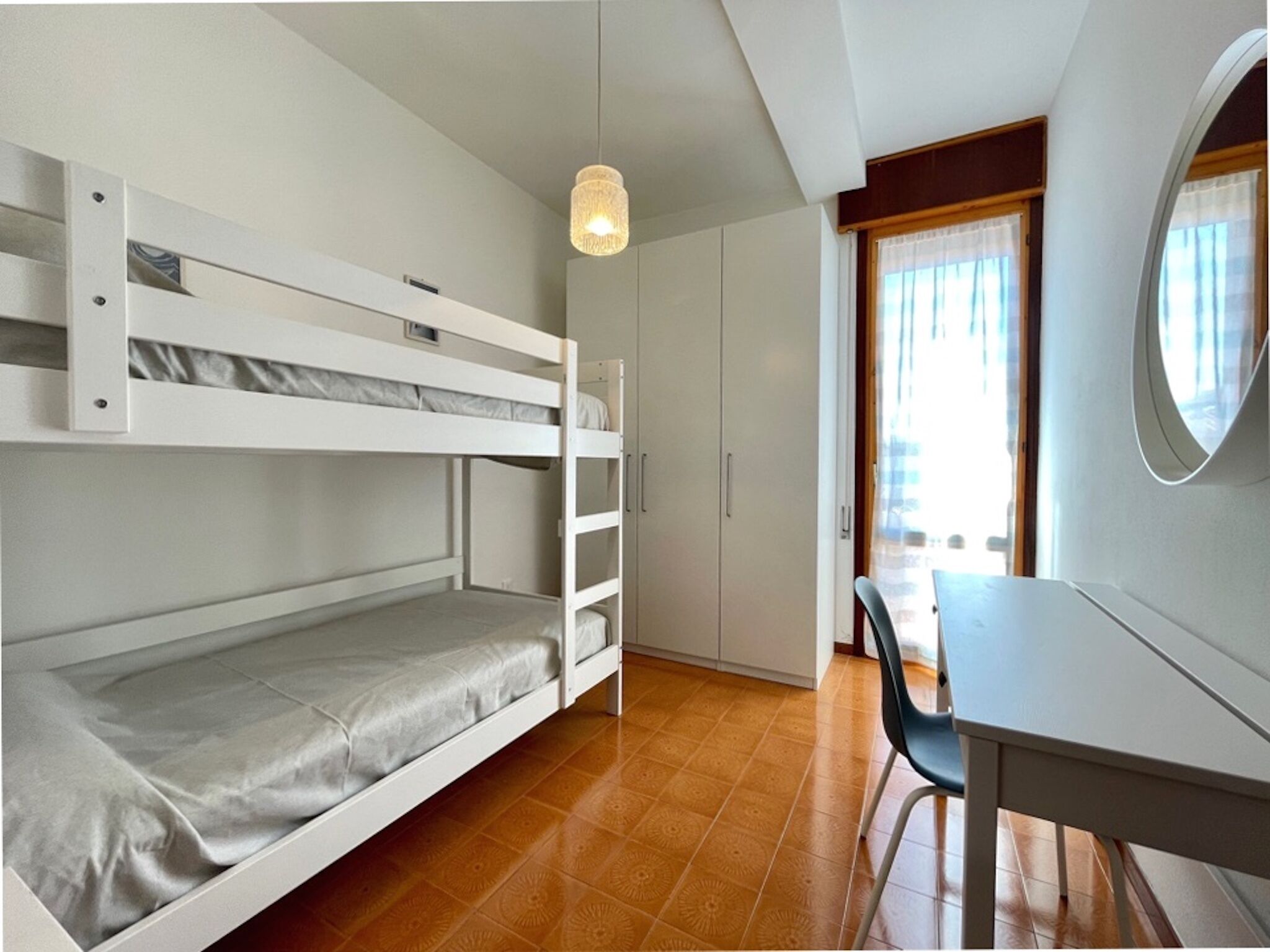 A bunk bed room with a desk and chair, a wardrobe, and a window with curtains.