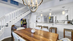 Un comedor amplio con una mesa de madera y sillas blancas, una escalera blanca y una cocina con electrodomésticos modernos.