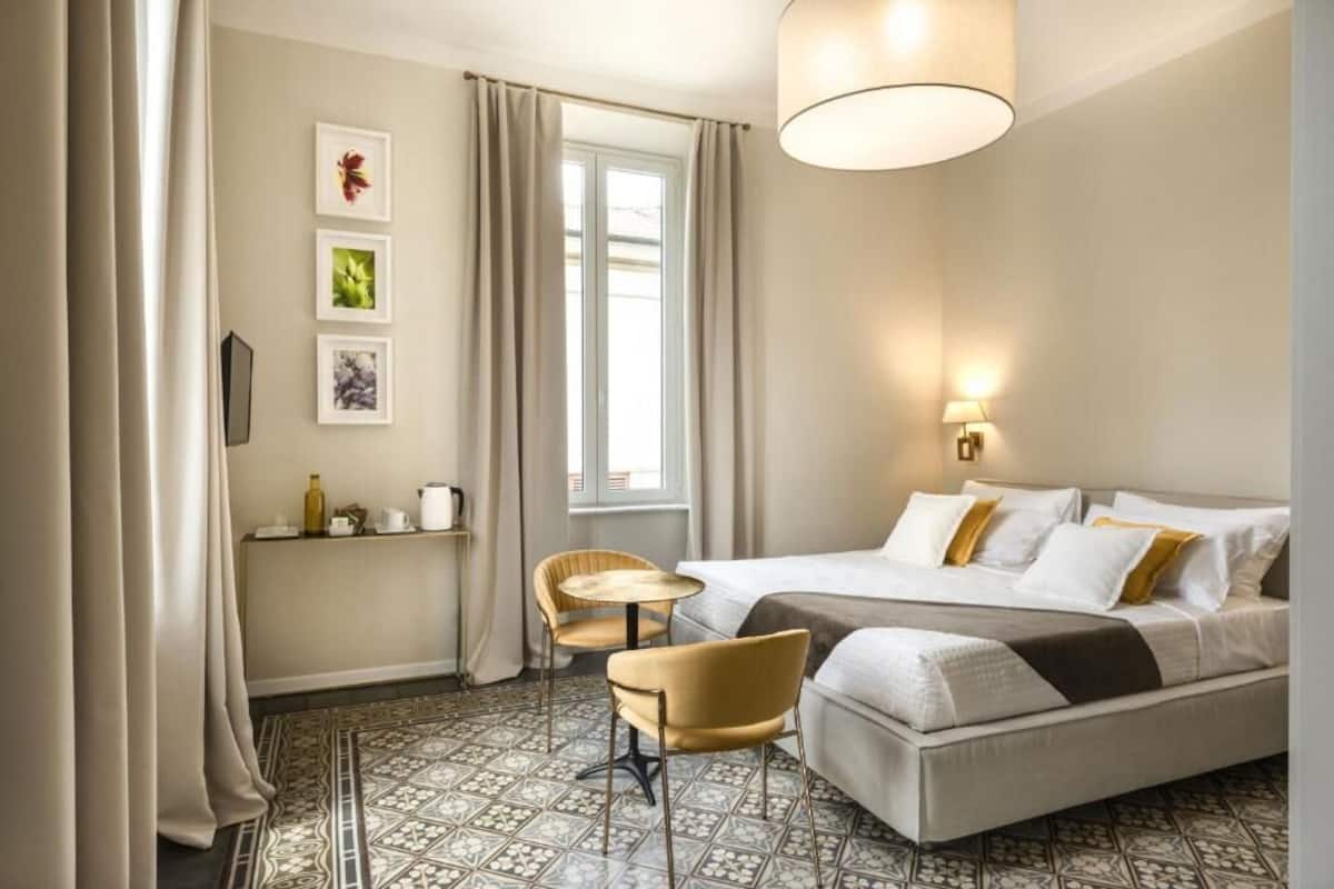 Habitación de hotel con cama, dos sillas, una mesita, ventana con cortinas y cuadros en la pared.