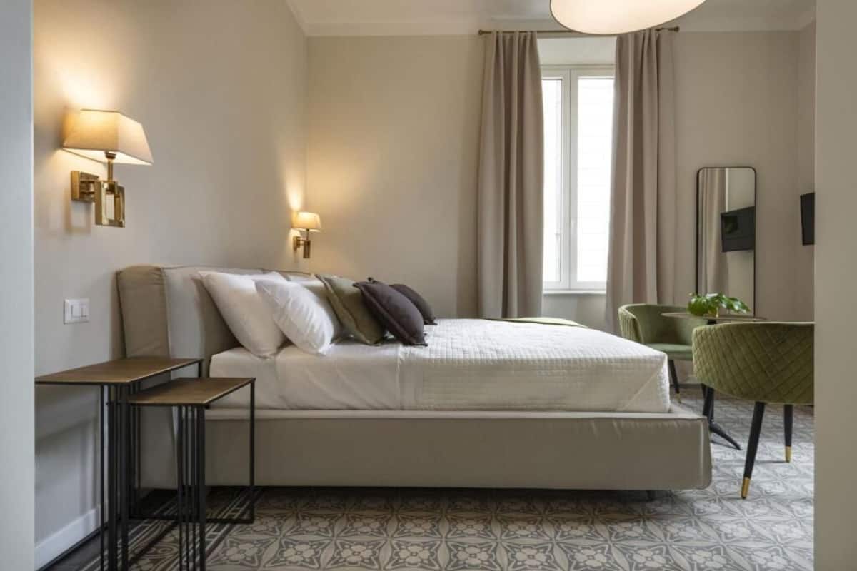 Un dormitorio con cama, mesitas de noche, lámpara, silla y ventana con cortinas.