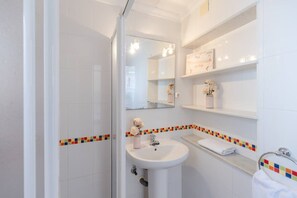 Ein Badezimmer mit Dusche, Waschbecken und Spiegel. Über dem Waschbecken befinden sich Regale mit Dekoration.
